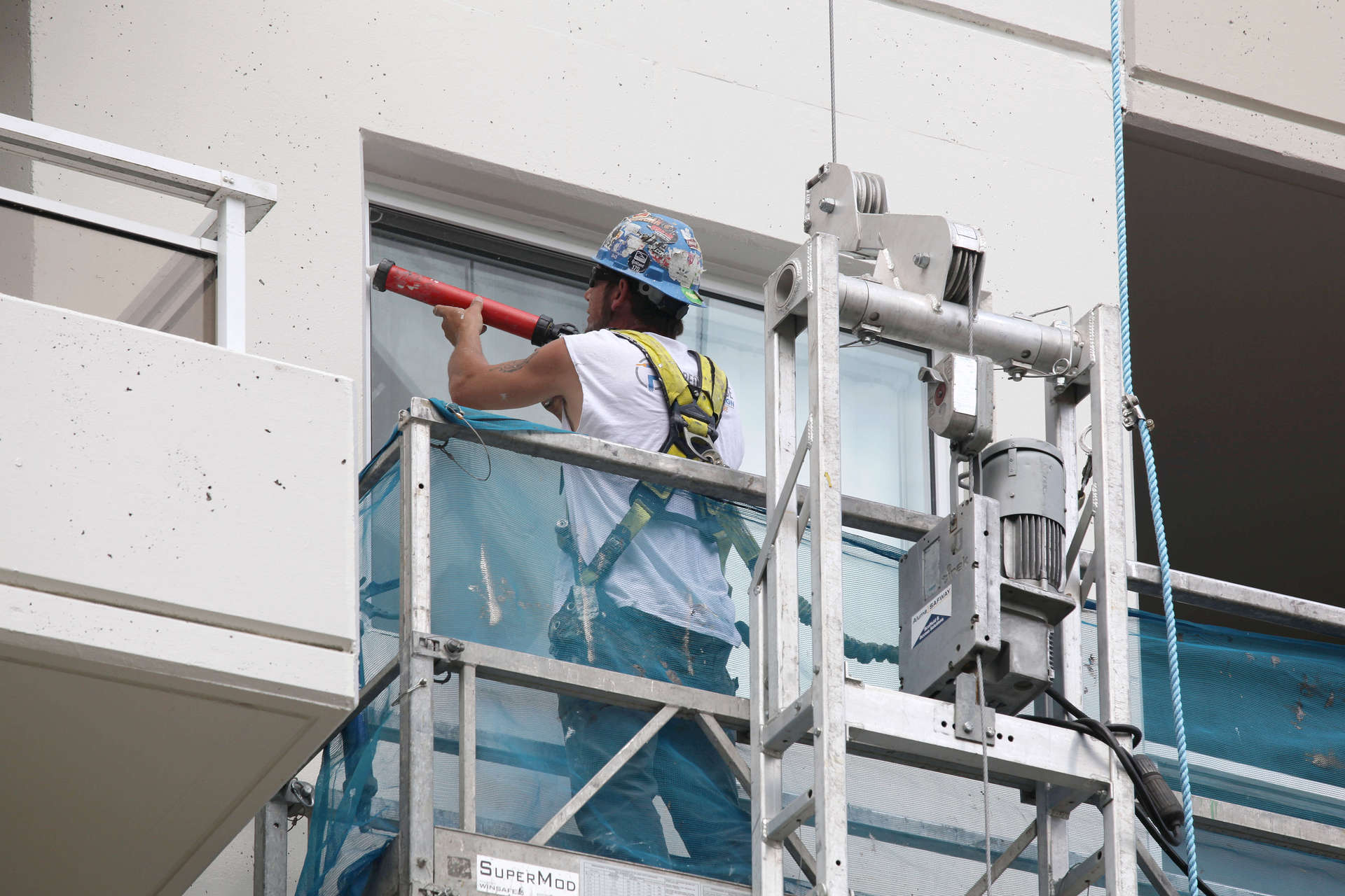 High rise building exterior painter with a caulking gun caulking windows on a weigh stage rig. A commercial and multi-unit residential high-rise exterior renovation, refresh, update - painting, caulking and coating. A high-rise painting project by Performance Painting and Caulking Ltd.